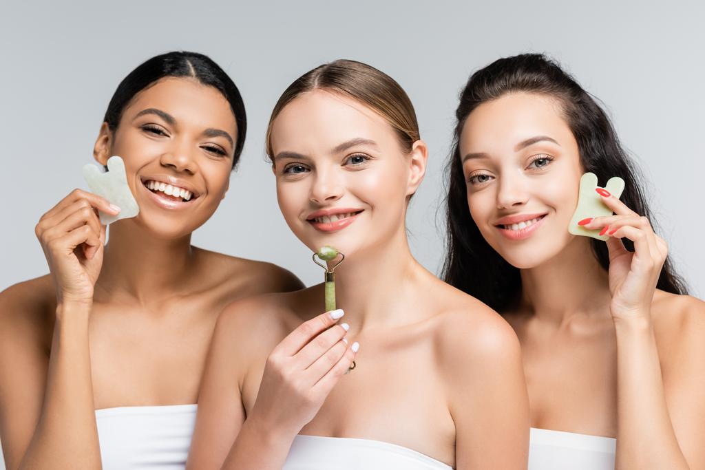 happy multiethnic women holding jade roller and gua sha facial tools isolated on grey - Photo, Image