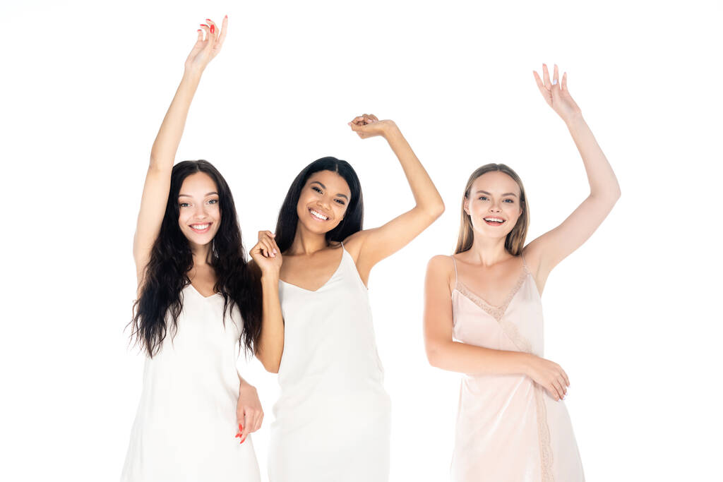 podekscytowane międzyrasowe kobiety w sukienki stojące z podniesionymi rękami odizolowane na białym - Zdjęcie, obraz