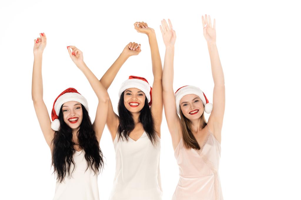 vrolijk interraciale vrouwen in santa hoeden en jurken staan met de handen boven hoofden geïsoleerd op wit - Foto, afbeelding
