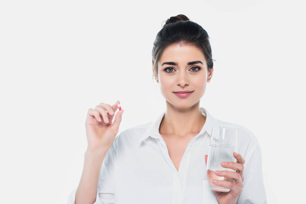 Brunette woman holding glass of water and pill while looking at camera isolated on white  - Photo, Image