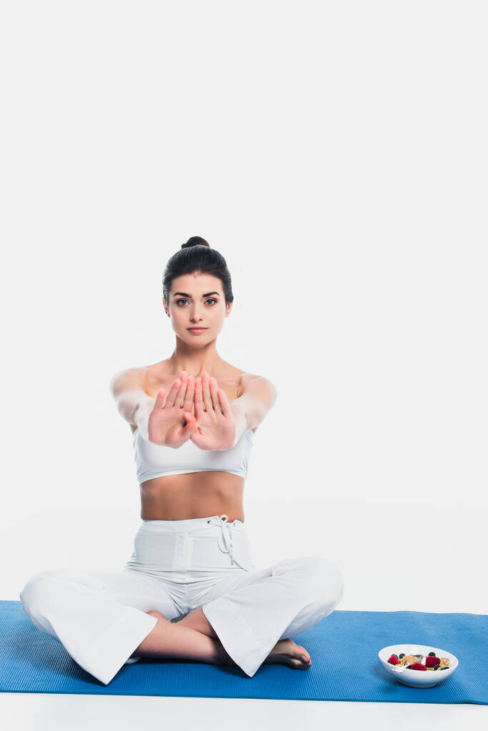 vrouw zitten in yoga pose in de buurt kom van granen met bessen op fitness mat op witte achtergrond  - Foto, afbeelding