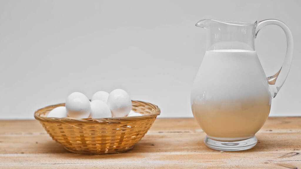 cesta de mimbre con un montón de huevos cerca de frasco de vidrio con leche fresca en la mesa de madera aislada en gris - Foto, imagen