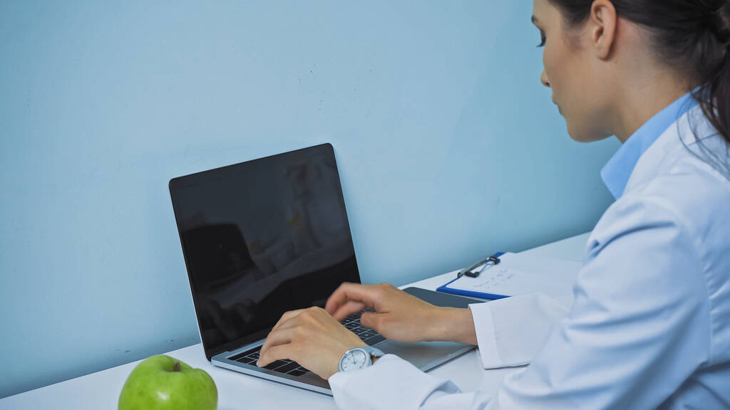 dentista escribiendo en el ordenador portátil con pantalla en blanco cerca de manzana fresca y portapapeles - Foto, Imagen
