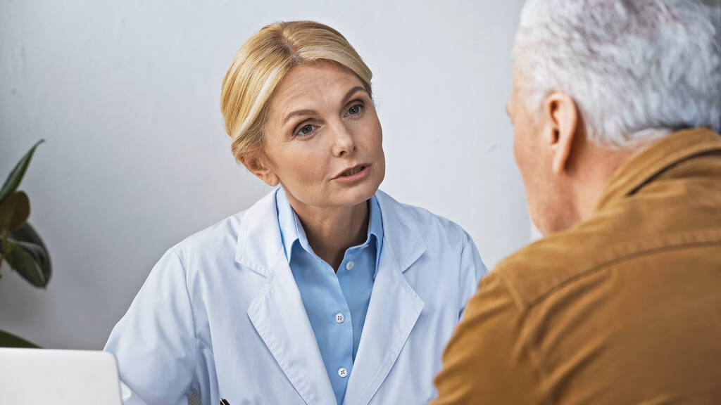 Dojrzały lekarz rozmawiający z pacjentem podczas wizyty w klinice, niewyraźny pierwszy plan - Zdjęcie, obraz