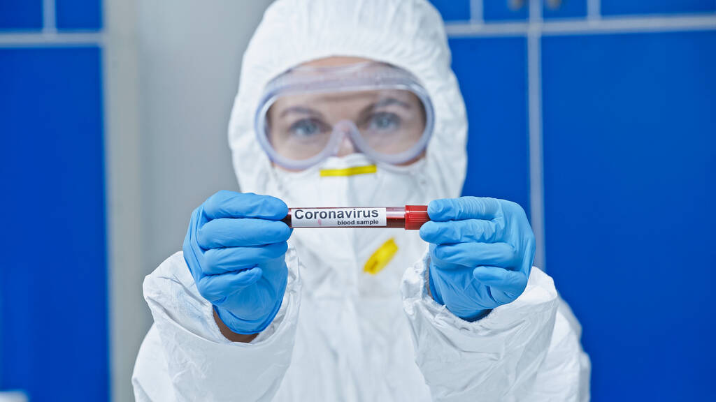 ιολόγος με στολή Hazmat που δείχνει δοκιμαστικό σωλήνα με γράμματα κορωναϊού στο εργαστήριο - Φωτογραφία, εικόνα