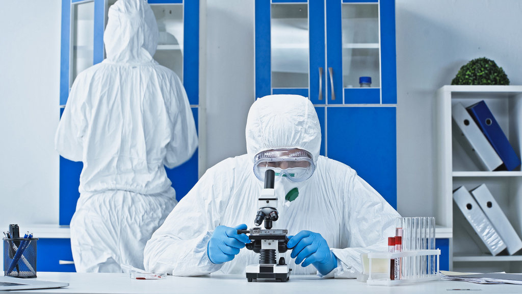 científico en traje de materiales peligrosos que trabaja con microscopio cerca de colega en laboratorio - Foto, imagen