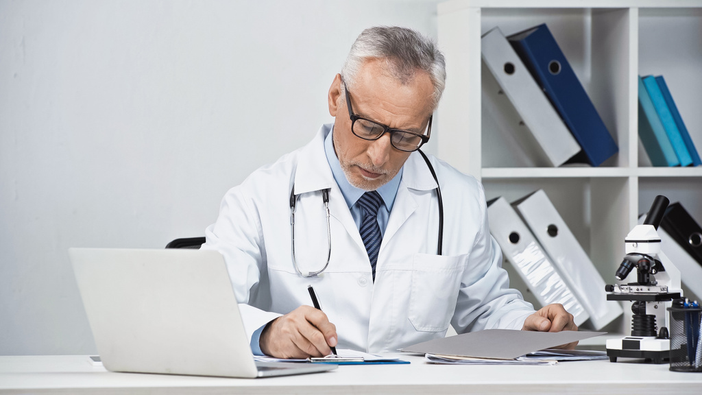 doctor en anteojos escribir prescripción cerca del ordenador portátil en el lugar de trabajo - Foto, Imagen