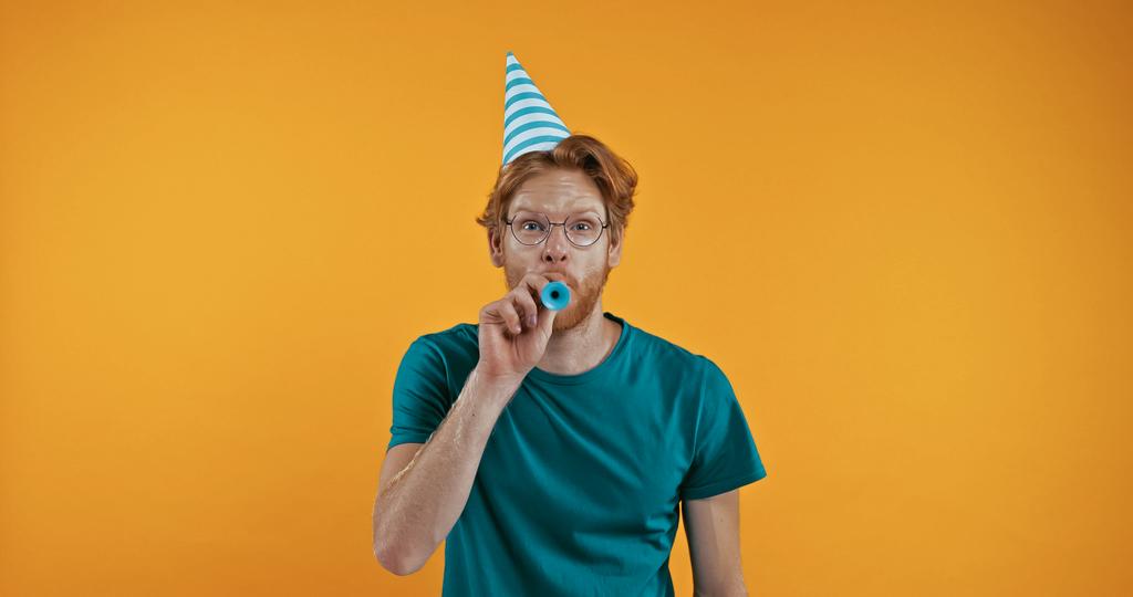 redhead man in party cap blowing in party horn isolated on yellow - Photo, Image