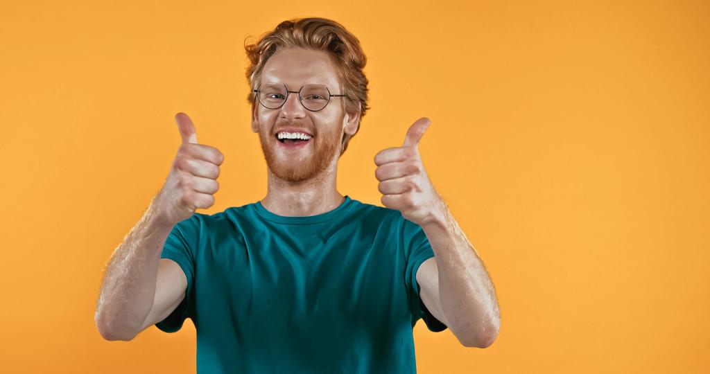 веселый рыжий мужчина в очках показывает большие пальцы вверх изолированы на желтый  - Фото, изображение