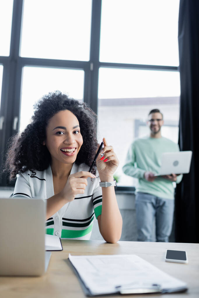 Femme d'affaires afro-américaine souriant à la caméra tout en tenant un stylo près d'un ordinateur portable et un presse-papiers sur le premier plan flou  - Photo, image