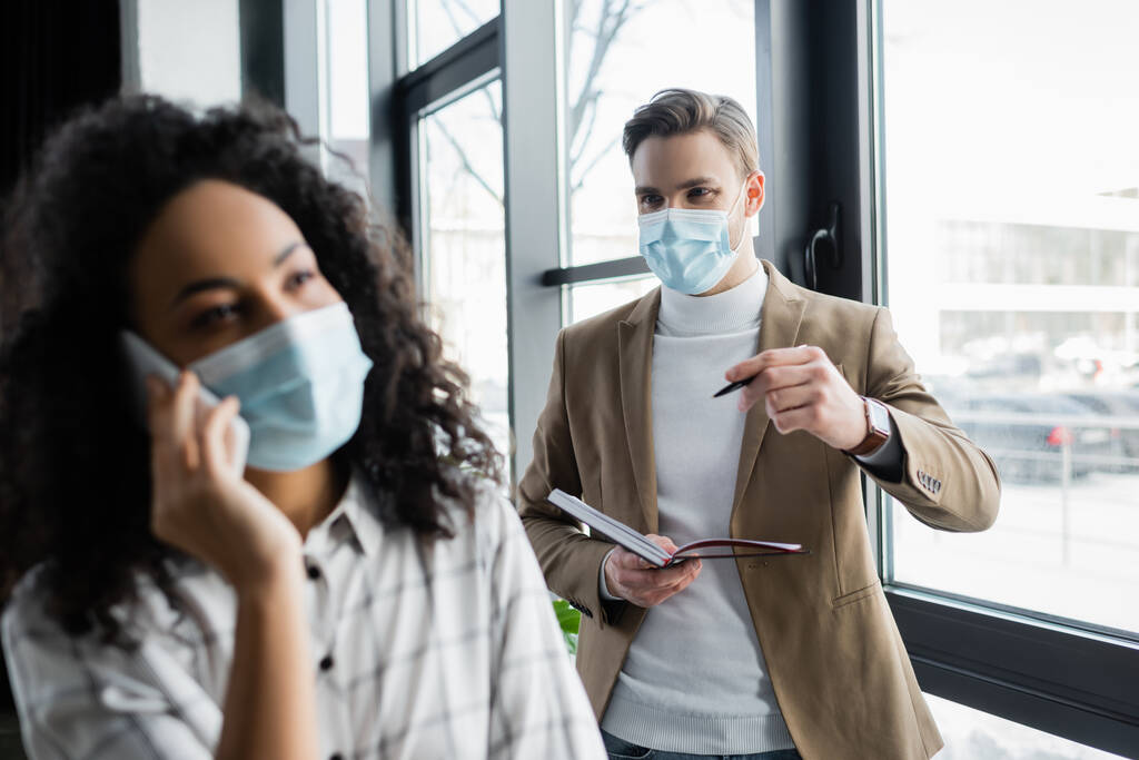 hombre de negocios en máscara médica con cuaderno y pluma cerca de colega afroamericano hablando en el teléfono inteligente, borrosa primer plano - Foto, imagen