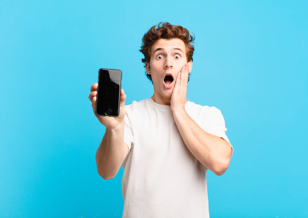 young boy feeling shocked and scared, looking terrified with open mouth and hands on cheeks. telephone screen concept - Photo, Image