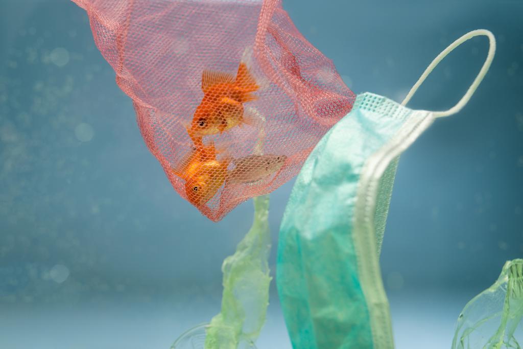 Plastikmüll, medizinische Maske und Netz mit Goldfischen im Wasser, ökologisches Konzept - Foto, Bild
