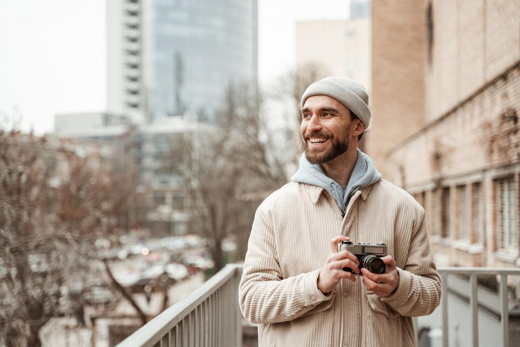 szakállas férfi sapkában és kabátban mosolyog, miközben retro kamerát tart  - Fotó, kép