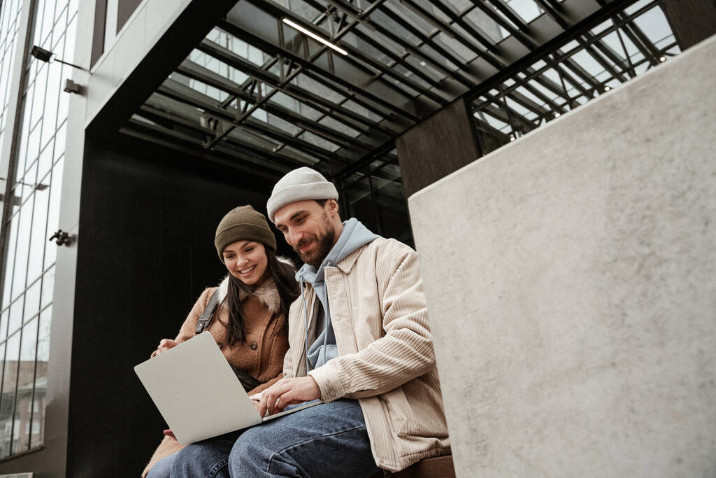 low angle view of freelancers in beanie hats smiling while using laptop - Photo, Image