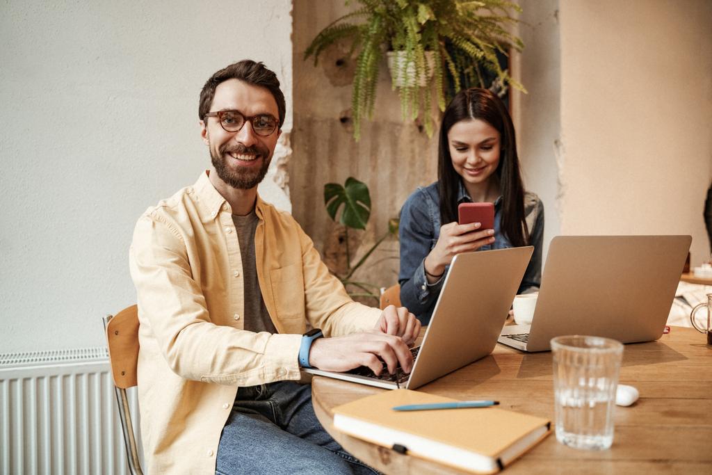 happy freelancer in eyeglasses using laptop near woman in cafe - Photo, Image
