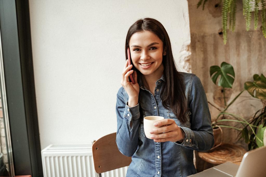 весела жінка говорить на смартфоні і тримає чашку кави
 - Фото, зображення