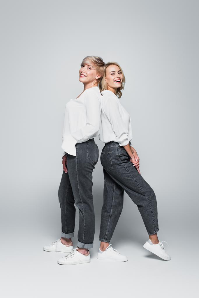 full length view of mother and daughter in trendy clothes standing back to back on grey - Photo, Image