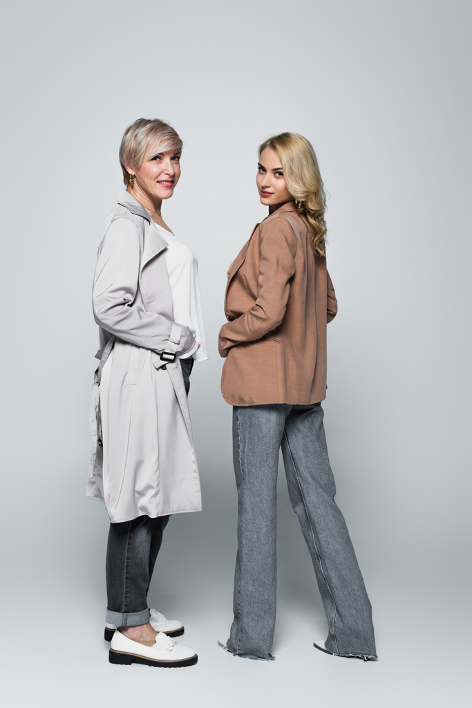 full length view of mature woman and adult daughter posing in trendy clothes on grey - Photo, Image