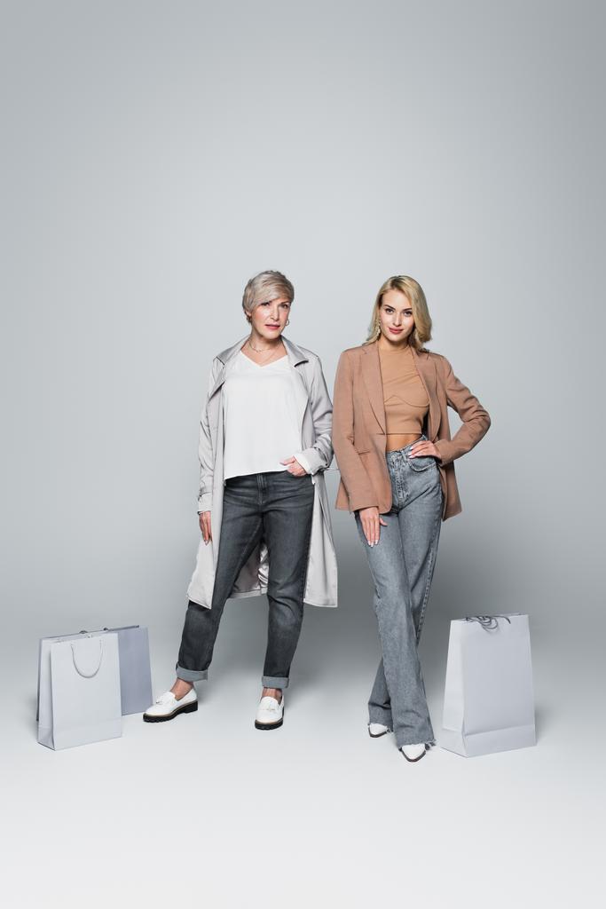 mother and daughter in trendy clothes posing near shopping bags on grey - 写真・画像