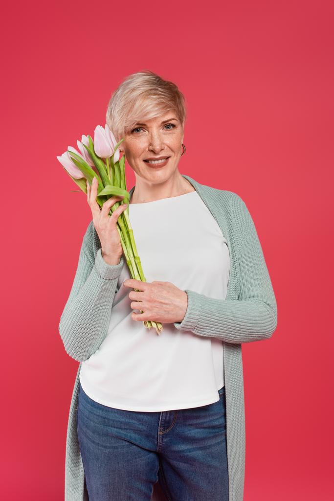 stylische Frau mittleren Alters lächelt in die Kamera, während sie Tulpen isoliert auf rosa hält - Foto, Bild