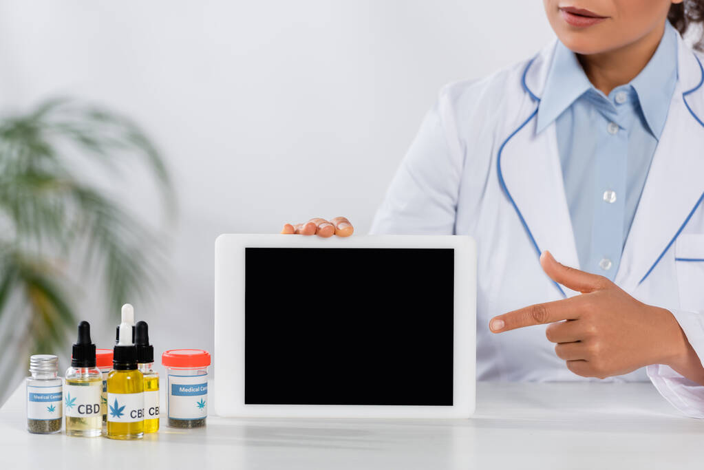 bijgesneden beeld van Afrikaanse Amerikaanse arts wijzend met de vinger naar digitale tablet met blanco scherm in de buurt van flessen met medische cannabis - Foto, afbeelding