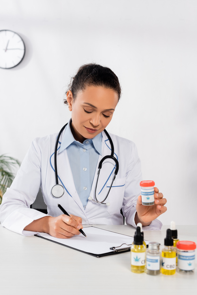 brunette médecin afro-américain avec stéthoscope tenant bouteille avec lettrage de cannabis médical et écriture sur presse-papiers - Photo, image