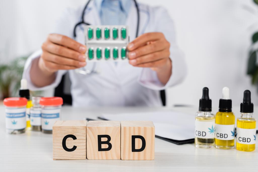 cubes with cbd lettering near doctor holding blister pack with capsules on blurred background - Photo, Image