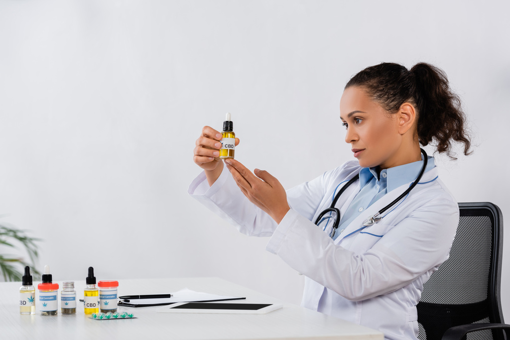 médico afroamericano sosteniendo botella de cbd cerca de la medicación y la tableta digital en el escritorio - Foto, Imagen