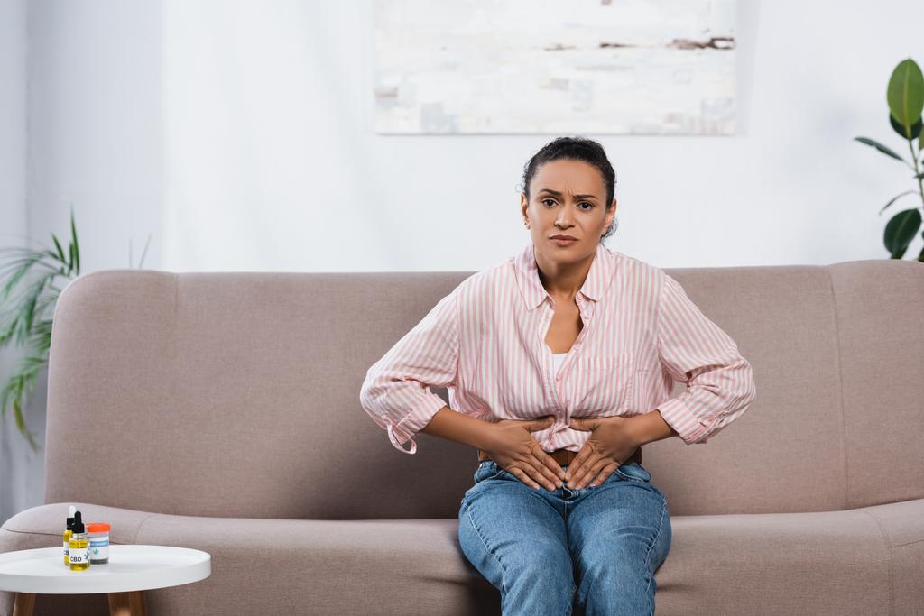 mujer afroamericana que sufre de dolor abdominal mientras se sienta en el sofá en la sala de estar  - Foto, Imagen