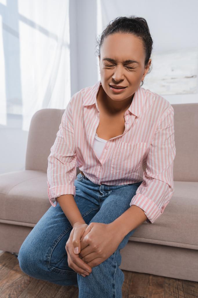 Afrikanische Amerikanerin leidet unter Schmerzen im Knie, während sie auf der Couch sitzt - Foto, Bild
