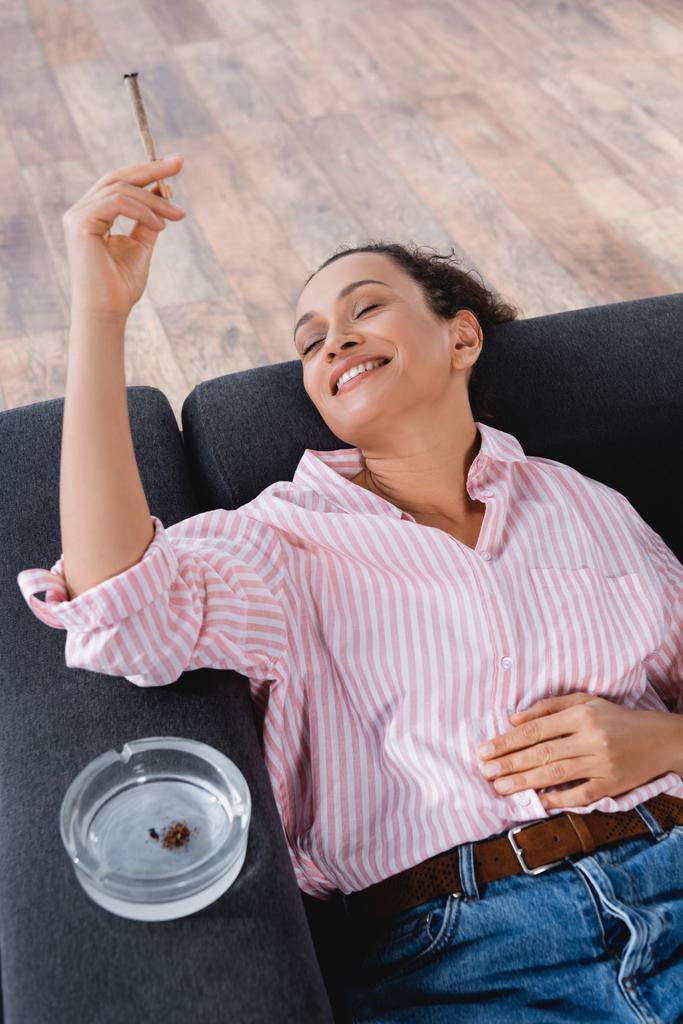 високий кут зору задоволеної афроамериканки лежить на дивані і курить спільно з медичним канабісом поблизу попільнички
  - Фото, зображення