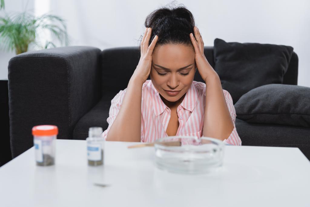 femme afro-américaine souffrant de migraine près de bouteilles de cannabis médical au premier plan flou - Photo, image