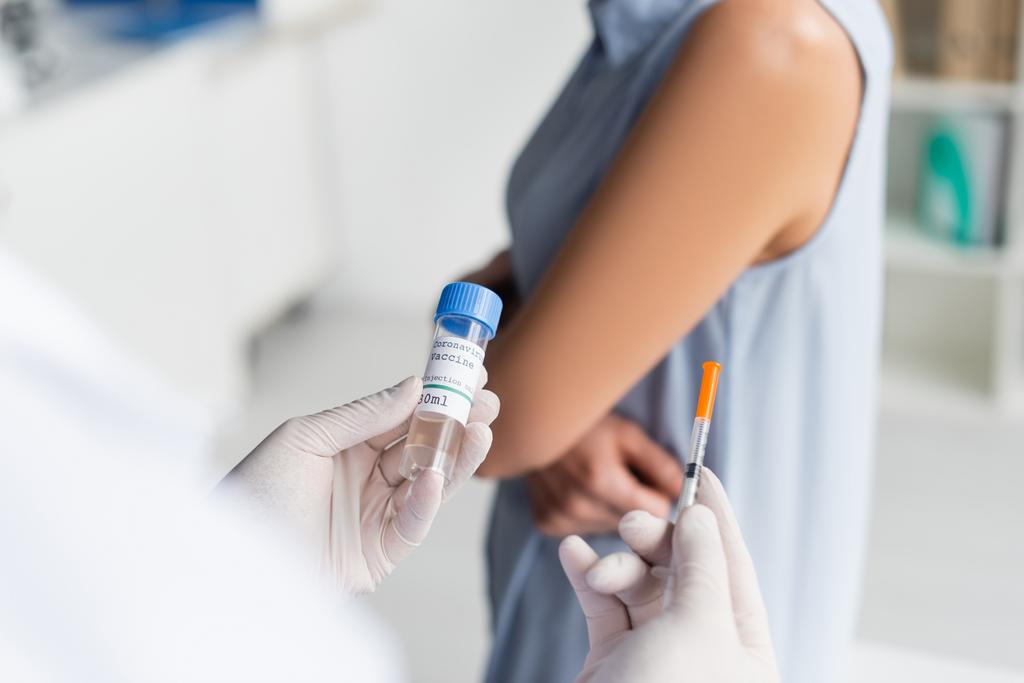 Ausgeschnittene Ansicht eines Arztes mit Coronavirus-Impfstoff und Spritze in der Nähe einer Frau auf verschwommenem Hintergrund  - Foto, Bild