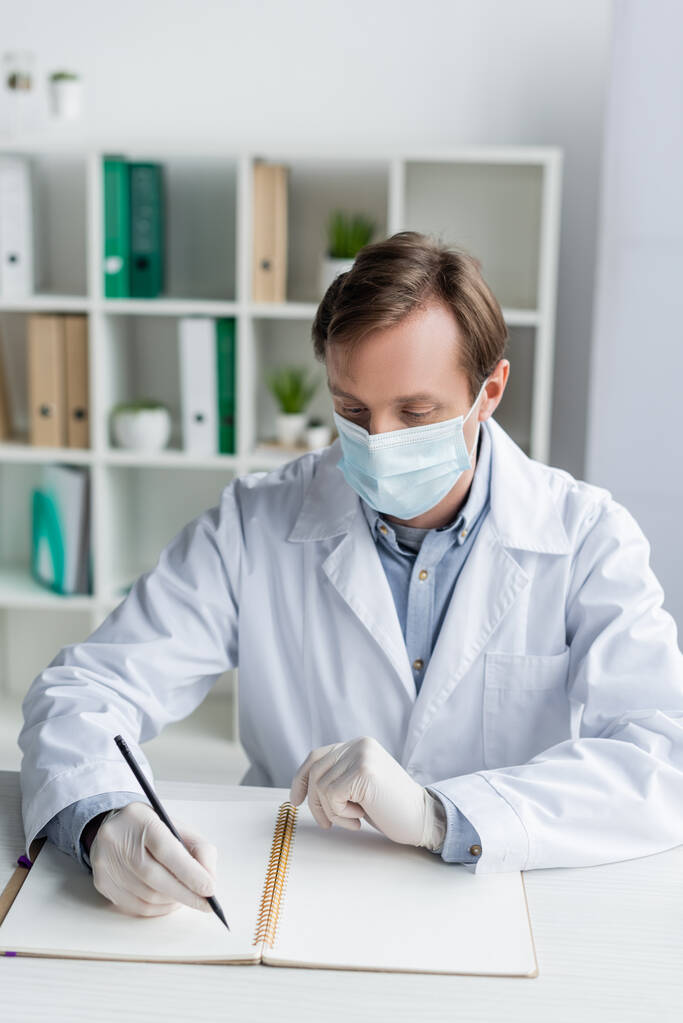 Лікар в медичній масці і латексні рукавички, що пишуть на блокноті за робочим столом
  - Фото, зображення