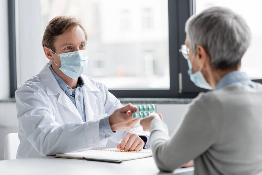 Врач в медицинской маске дает пациенту таблетки на размытом переднем плане в клинике  - Фото, изображение