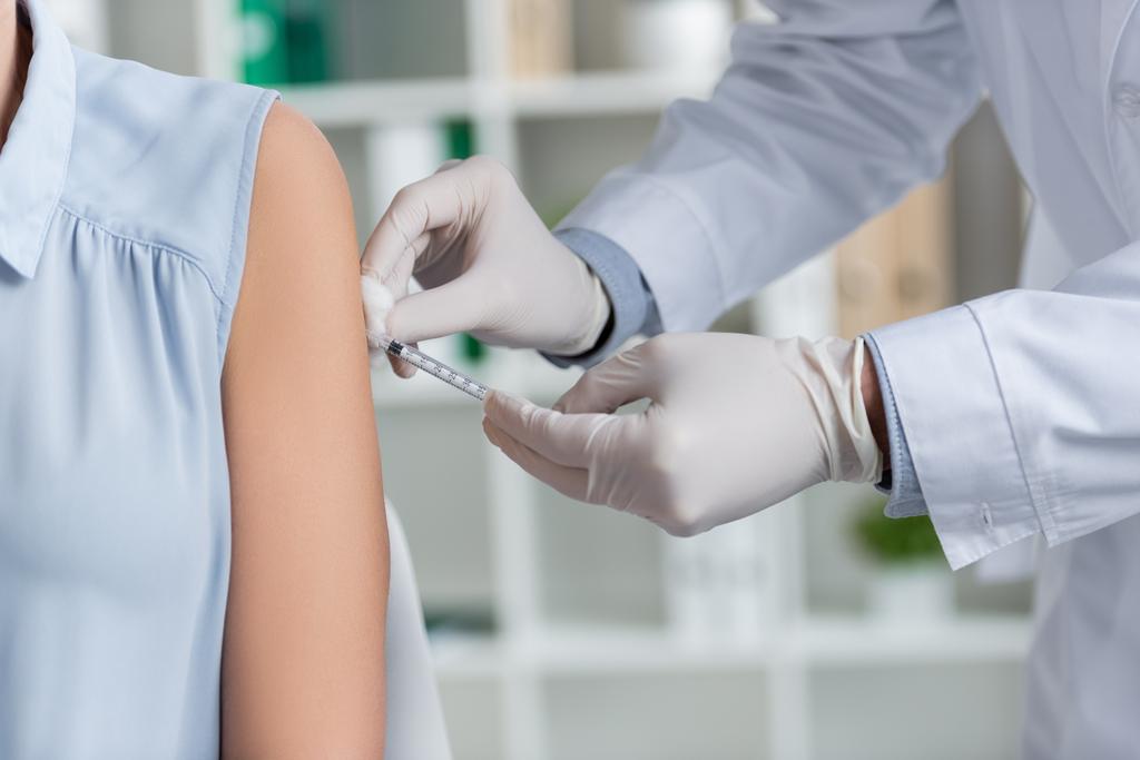 Oříznutý pohled na lékaře v latexových rukavicích držících injekční stříkačku a provádějících očkování ženy v nemocnici  - Fotografie, Obrázek
