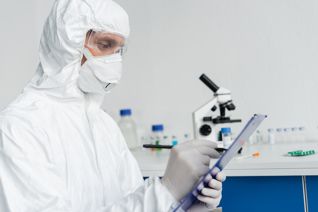 Scientist in protective uniform writing on clipboard on blurred foreground  - Photo, Image