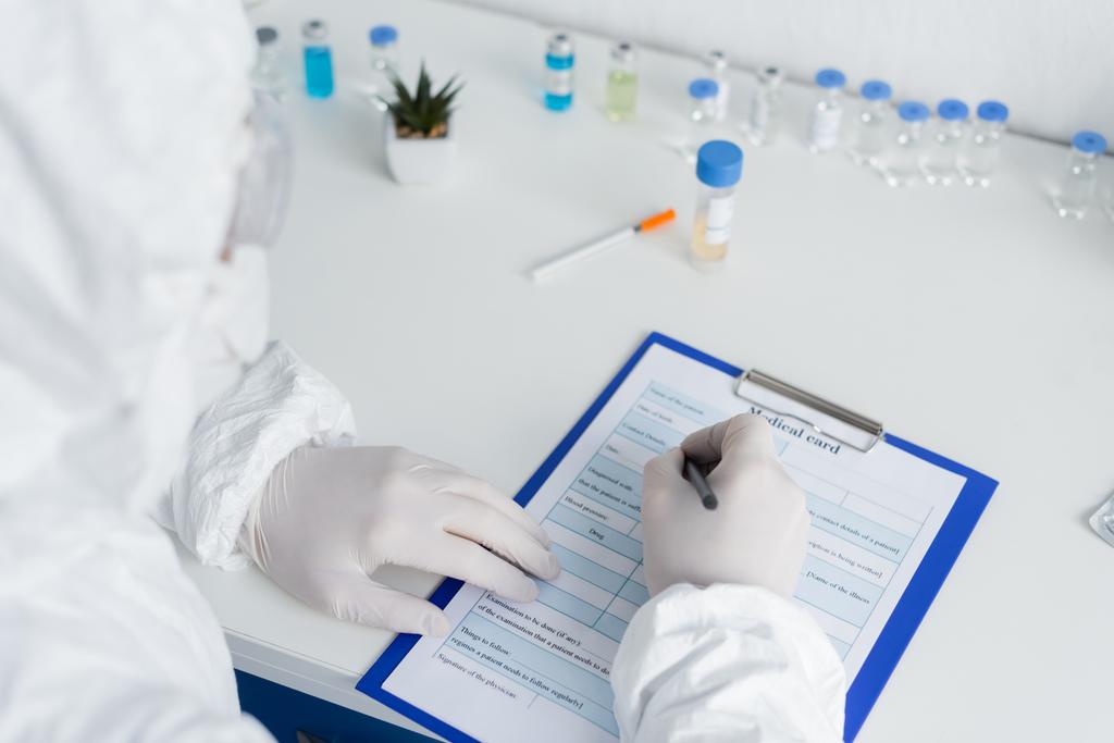 Cientista em primeiro plano desfocado escrevendo no cartão médico perto de vacinas em laboratório  - Foto, Imagem