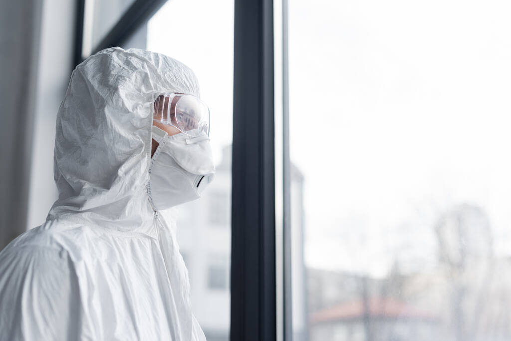 Científico en máscara médica, gafas y traje de materiales peligrosos de pie cerca de la ventana  - Foto, Imagen