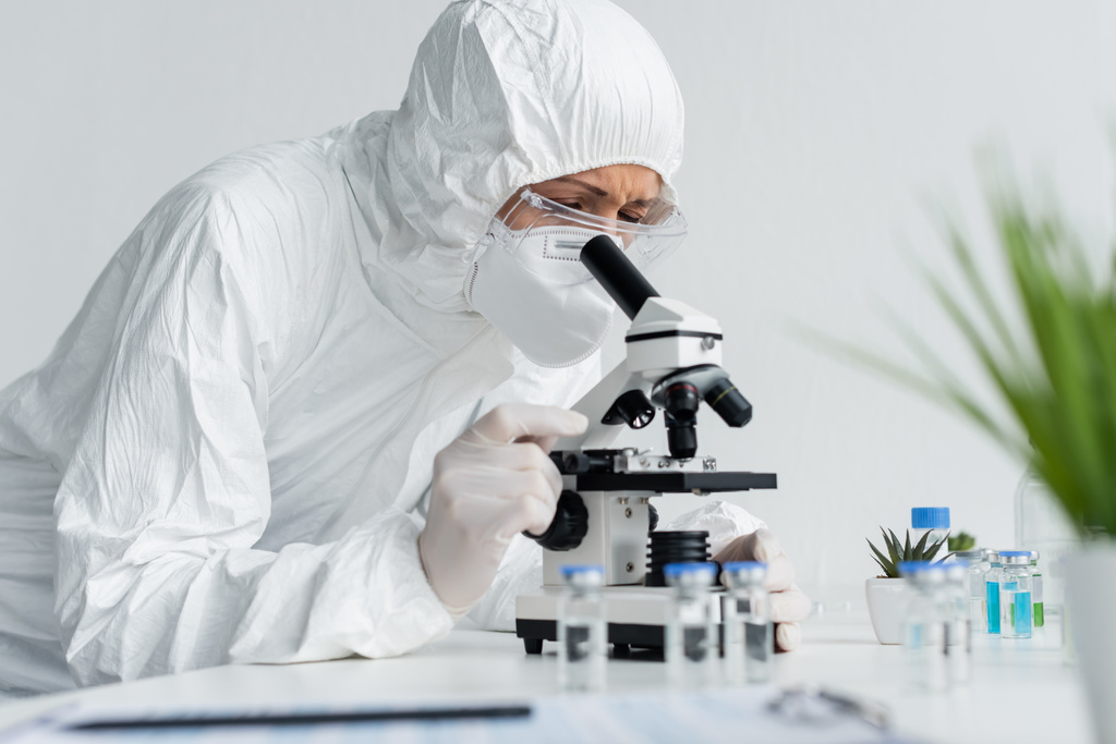 Tehlikeli madde giysisi giyen bilim adamları bulanık ön planda aşıların yanında mikroskop ile çalışıyorlar.  - Fotoğraf, Görsel