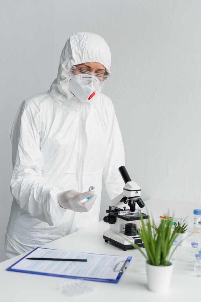 Ученый в защитном костюме рассматривает вакцину возле буфера обмена и микроскопа в лаборатории  - Фото, изображение