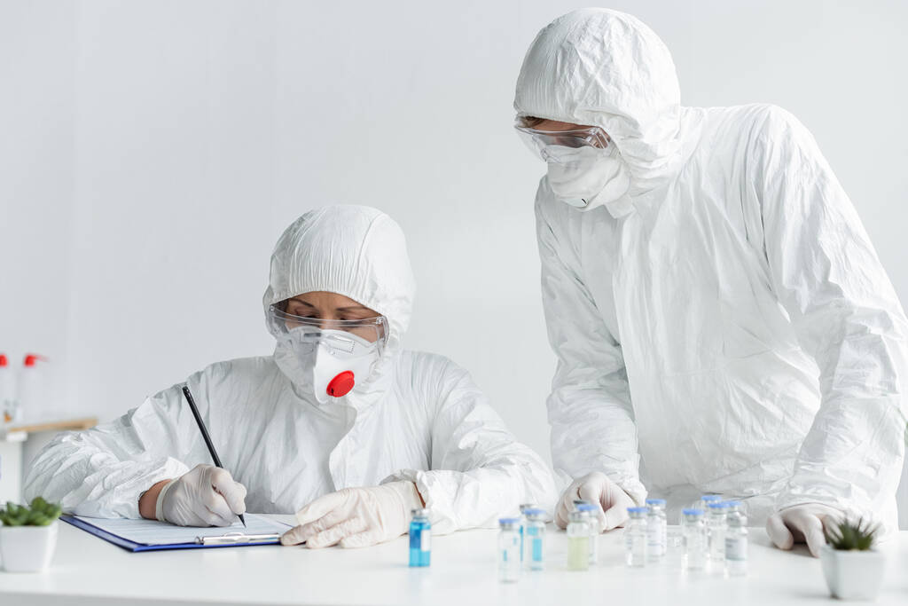Ученый пишет в буфер обмена рядом с коллегой и вакцины на размытом переднем плане  - Фото, изображение