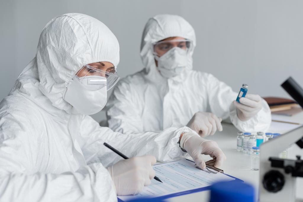 Científico escribiendo en portapapeles cerca de colega con vacunas sobre fondo borroso  - Foto, Imagen