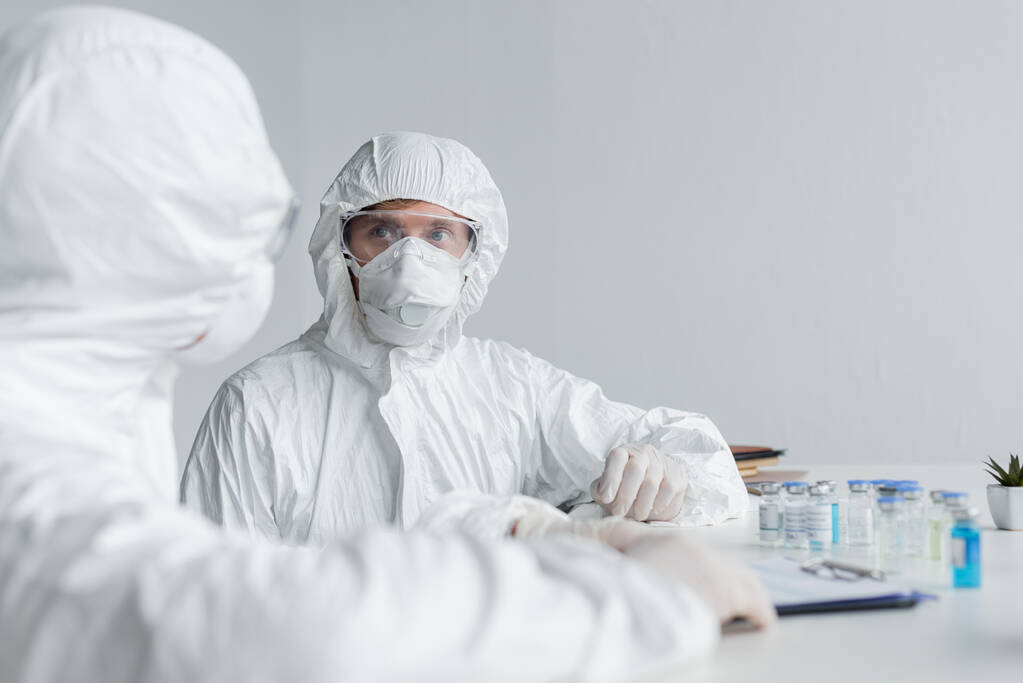 Wissenschaftler im Warnanzug schaut Kollege in der Nähe von Klemmbrett und Impfstoffen an  - Foto, Bild