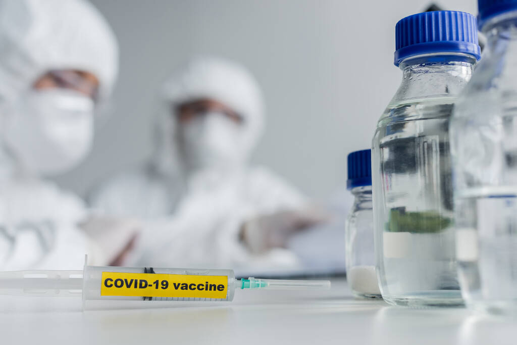 Spritze mit Covid-19 Impfstoff-Schriftzug in der Nähe von Wissenschaftlern im Labor  - Foto, Bild