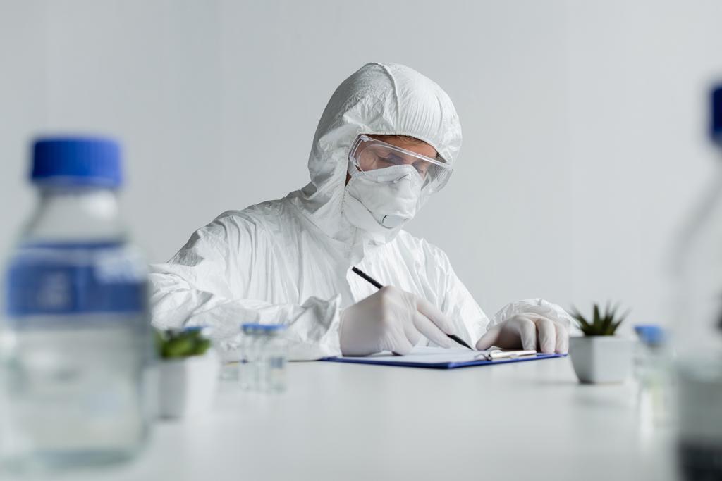 Scientist writing on clipboard near vaccines and plants on blurred foreground  - Photo, Image