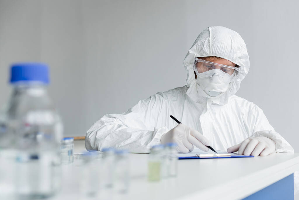 Scientist writing on clipboard near vaccines on blurred foreground on table  - Photo, Image