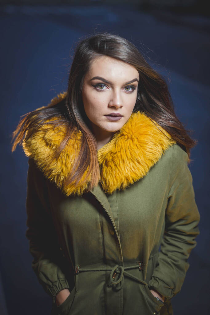 Retrato vertical de una joven atractiva con un abrigo de piel amarilla posando en una calle por la noche - Foto, Imagen
