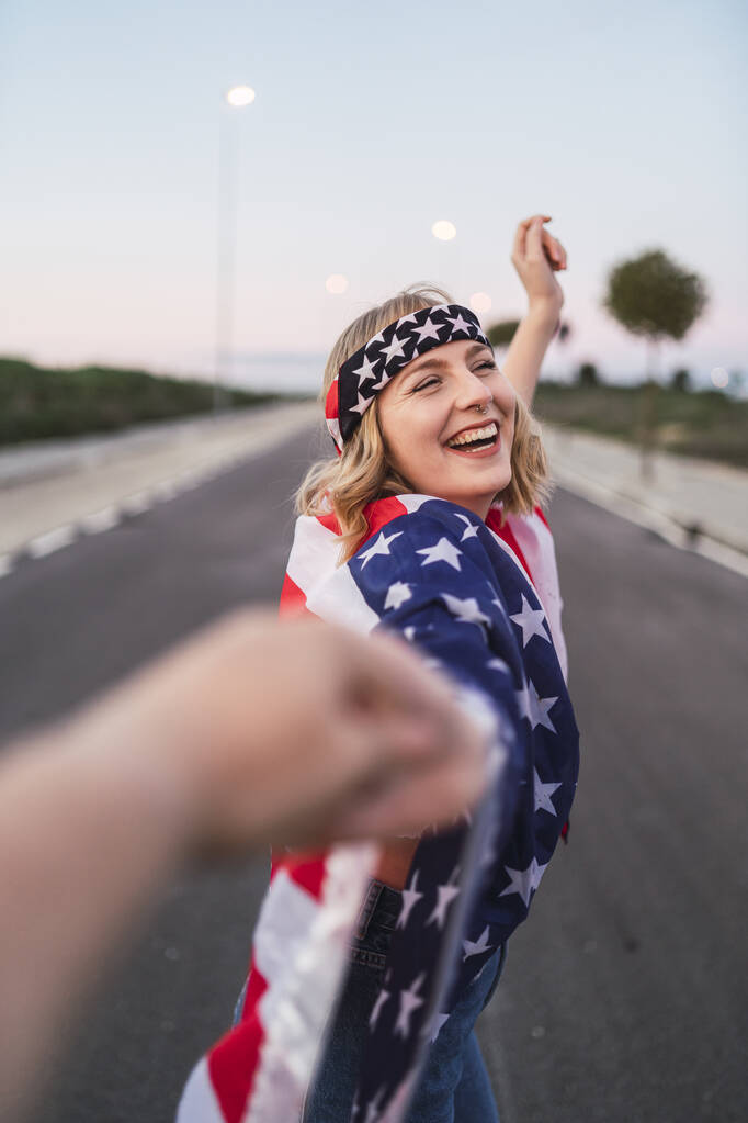 Una mujer caucásica feliz con el pelo corto y rubio y una bandera de Estados Unidos sosteniendo una mano mientras camina por el camino - Foto, imagen